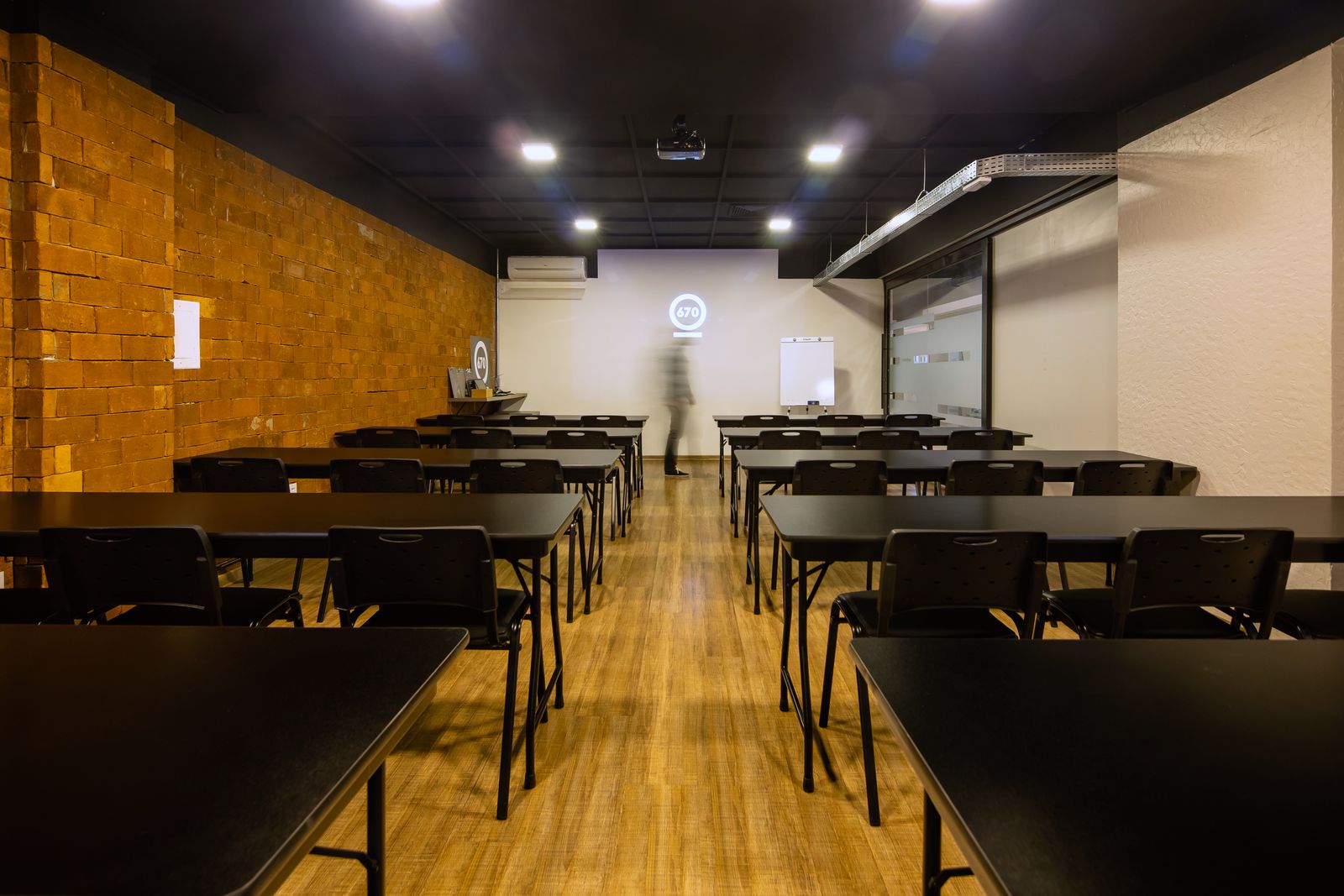 Sala Multiuso montada em formato de sala de aula, com mesas pretas, parede de tijolos e projetor