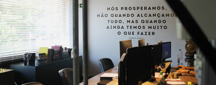 Fotografia ilustrativa de um espaço do coworking e startups. Na parede, tem a frase "nós prosperamos, não quando alcançamos tudo, mas quando ainda temos muito o que fazer"